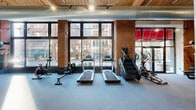 This fitness center combines industrial-chic architectural character with a full suite of exercise equipment, creating a functional and visually engaging workout environment. The space spans a generous single floor, thoughtfully divided into distinct training zones to accommodate a variety of fitness needs.

The cardio area is a standout feature, positioned directly in front of expansive floor-to-ceiling windows that frame street-level views of the surrounding urban neighborhood. Treadmills, an elliptical, a stair climber, and a stationary bike line this naturally lit zone, benefiting from abundant daylight that energizes the space throughout the day.

The strength training area occupies the rear of the facility, featuring a power rack, a cable machine, a multi-purpose bench, dumbbells, kettlebells, medicine balls, and a rowing machine. Full-length mirrors line one wall, enhancing both functionality and the sense of openness.

Architecturally, the space is defined by exposed brick walls, warm wood-panel ceiling accents, and prominent industrial-style ductwork overhead. Track lighting supplements natural light throughout, ensuring consistent illumination across all training zones. Durable rubber and carpet flooring provide safe, supportive surfaces underfoot. The street-facing glass entrance adds transparency and connectivity to the active urban setting outside, contributing to the space's energetic and welcoming ambiance.