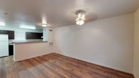 This two-floor space offers a clean, functional layout with notable design details throughout. The main living area is highlighted by rich, dark hardwood flooring that runs continuously across the room, adding warmth and a sense of cohesion to the space. White walls provide a neutral backdrop, while a ceiling fan with a light fixture serves as a practical central feature overhead. A textured ceiling adds subtle visual interest above.

A floor-mounted HVAC vent is integrated into the wall, ensuring climate control throughout the space. The living area flows naturally toward a kitchen, partially visible in the background, which features dark cabinetry and a countertop pass-through or bar area, hinting at an open-concept design between the two spaces. Recessed and flush-mount lighting illuminate both areas effectively.

Multiple electrical outlets are distributed along the walls, offering practical convenience throughout. The overall ambiance is modest and move-in ready, with a straightforward layout that balances open living space with functional utility. The two-floor configuration suggests additional living or sleeping areas on the upper level, providing separation between common and private spaces.