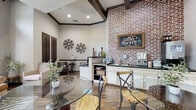 This inviting single-floor communal space blends rustic charm with contemporary design to create a warm and functional gathering environment. The centerpiece of the space is a dramatic floor-to-ceiling exposed brick accent wall, which anchors a well-appointed coffee bar — complete with dark granite countertops, white cabinetry, a sink, and a coffee machine beneath a charming chalkboard sign reading "Coffee Therapy." Adjacent to the bar, a reception desk with a computer workstation provides a practical touch, while a wall-mounted television is neatly recessed into a built-in niche nearby.

The main lounge area features soaring vaulted ceilings with striking dark wood exposed beams, contributing to an airy, loft-like atmosphere. Rich hardwood flooring runs throughout, adding warmth and continuity to the space. A round glass-top dining table with metal and wood bistro-style chairs offers a casual seating arrangement, while a plush leather sofa and neutral upholstered armchairs — set atop a bold zebra-print area rug — create a relaxed conversational seating zone.

Tall windows dressed with patterned sheer curtains invite abundant natural light and offer views of the surrounding greenery outside. Decorative accents, including vintage-style windmill wall art, ceramic vases, and potted greenery, complete the eclectic yet cohesive aesthetic of this thoughtfully designed communal retreat.