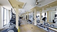 This fitness center offers a thoughtfully designed workout environment that combines functionality with refined aesthetics. The space features a coffered ceiling with recessed lighting and multiple ceiling fans, contributing to a well-ventilated and visually appealing atmosphere. Structural columns are positioned throughout the floor, adding an architectural rhythm to the layout while helping to define distinct workout zones. The flooring consists of a multi-toned carpet with subtle yellow and gray accents that complement the neutral, cream-toned walls and columns. A generous selection of cardio equipment is prominently displayed, including StairMaster steppers, elliptical trainers, treadmills, and rowing machines, all arranged in organized rows to maximize the available floor space. Strength training equipment and weight machines are also visible toward the rear of the space, accommodating a full range of fitness routines. Large mirrors line the walls, enhancing the sense of openness and allowing users to monitor their form during workouts. Natural light filters in through windows visible at the far end, further brightening the interior. A flat-screen television is mounted for entertainment during workouts, rounding out the amenities of this comprehensive and well-appointed fitness facility.