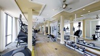 This fitness center offers a thoughtfully designed workout environment that combines functionality with refined aesthetics. The space features a coffered ceiling with recessed lighting and multiple ceiling fans, contributing to a well-ventilated and visually appealing atmosphere. Structural columns are positioned throughout the floor, adding an architectural rhythm to the layout while helping to define distinct workout zones. The flooring consists of a multi-toned carpet with subtle yellow and gray accents that complement the neutral, cream-toned walls and columns. A generous selection of cardio equipment is prominently displayed, including StairMaster steppers, elliptical trainers, treadmills, and rowing machines, all arranged in organized rows to maximize the available floor space. Strength training equipment and weight machines are also visible toward the rear of the space, accommodating a full range of fitness routines. Large mirrors line the walls, enhancing the sense of openness and allowing users to monitor their form during workouts. Natural light filters in through windows visible at the far end, further brightening the interior. A flat-screen television is mounted for entertainment during workouts, rounding out the amenities of this comprehensive and well-appointed fitness facility.