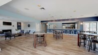 Located at 1 Makefield Rd in Morrisville, PA, this single-floor community clubhouse offers a well-rounded amenity space designed for both relaxation and recreation. The main gathering area is an open, airy room finished with light hardwood flooring and recessed lighting throughout. A bold navy blue bar counter with bar-height seating runs along one wall beneath a decorative "gather" sign, opening directly into a fully equipped kitchen featuring stainless steel appliances, cabinetry, and a tile backsplash. Multiple high-top tables and chairs fill the space, making it ideal for social events and casual gatherings. Sliding glass doors allow natural light to pour in, enhancing the bright, welcoming atmosphere. Adjacent to the main hall is a cozy lounge area anchored by a classic fireplace framed with white built-in shelving and cabinetry. A wall-mounted television sits above the mantle, while comfortable seating and a coffee table complete this inviting retreat. A dedicated billiards room offers a full-size pool table set atop plush carpeting, surrounded by large windows with ample natural light. Completing the clubhouse is a curved fitness center equipped with ellipticals, stationary bikes, and a treadmill, with track lighting and interior glass windows providing an open, energetic environment.