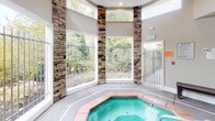 Located in Puyallup, WA, this single-floor indoor spa and wellness facility offers a thoughtfully designed retreat centered around relaxation and comfort. The heart of the space is a large octagonal hot tub, framed by terracotta-tiled surrounds and fitted with stainless steel entry rails for easy access. The pool area benefits from soaring ceilings and generous natural light streaming through tall windows, which are accented by striking stacked-stone columns that lend a warm, lodge-like aesthetic to the room. Lush greenery is visible through the windows, adding a connection to the outdoors. Adjacent to the hot tub is a classic wood-paneled sauna, fully lined in rich cedar from floor to ceiling — including tiered benches and a slatted ceiling — creating an immersive, traditional sauna experience. A rinse shower station with a tiled surround is conveniently positioned nearby. The main area also includes a seating bench for relaxation and coat hooks for personal belongings. Durable aggregate flooring flows throughout, while high clerestory windows enhance the sense of openness. The facility is well-equipped with safety features including an emergency call station, fire alarm, and clearly marked exits.