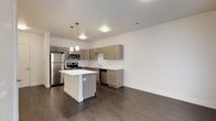 This contemporary single-floor residence showcases a thoughtfully designed open-concept layout that seamlessly connects the kitchen and living areas. The kitchen is a standout feature, equipped with stainless steel appliances including a refrigerator, range, microwave, and dishwasher. Textured wood-grain cabinetry lines the walls in an L-shaped configuration, complemented by a classic white subway tile backsplash and light-toned countertops. A central kitchen island with a white surface provides additional workspace and casual seating. Stylish pendant lights hang above the island, adding a modern touch to the space. Dark hardwood-style flooring runs consistently throughout, grounding the open layout with warmth and cohesion. The living and entry areas are bright and clean, with crisp white walls, recessed lighting, and a glass-panel door that invites natural light. A doorway leads to a carpeted bedroom, offering a soft contrast to the hard flooring in the main living spaces. The bathroom continues the home's modern aesthetic, featuring wood-grain cabinetry, a white countertop with an integrated sink, a large mirror, and a full tub-shower combination. The overall design is clean, functional, and contemporary throughout.