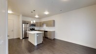 This contemporary single-floor residence showcases a thoughtfully designed open-concept layout that seamlessly connects the kitchen and living areas. The kitchen is a standout feature, equipped with stainless steel appliances including a refrigerator, range, microwave, and dishwasher. Textured wood-grain cabinetry lines the walls in an L-shaped configuration, complemented by a classic white subway tile backsplash and light-toned countertops. A central kitchen island with a white surface provides additional workspace and casual seating. Stylish pendant lights hang above the island, adding a modern touch to the space. Dark hardwood-style flooring runs consistently throughout, grounding the open layout with warmth and cohesion. The living and entry areas are bright and clean, with crisp white walls, recessed lighting, and a glass-panel door that invites natural light. A doorway leads to a carpeted bedroom, offering a soft contrast to the hard flooring in the main living spaces. The bathroom continues the home's modern aesthetic, featuring wood-grain cabinetry, a white countertop with an integrated sink, a large mirror, and a full tub-shower combination. The overall design is clean, functional, and contemporary throughout.
