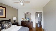 This two-floor residence offers a comfortable and well-designed living environment. The bedroom showcases warm hardwood flooring that adds richness and character to the space. Neutral-toned walls in soft white and greige tones create a calm, restful atmosphere, complemented by smooth white ceilings fitted with a ceiling fan featuring a central light fixture — providing both comfort and ambient lighting year-round.

The bedroom is spacious enough to accommodate a full furniture arrangement, including a bed with a classic headboard, a dark-stained wood dresser topped with a framed mirror, and matching nightstands with table lamps. The dark wood furniture provides an elegant contrast against the light walls, lending a traditional yet timeless aesthetic to the room.

A doorway off the bedroom leads directly into an en-suite bathroom, which features tiled flooring and appears to include standard bath fixtures. Natural light filters through a window in the bathroom, brightening the adjoining space. The overall ambiance of the bedroom is serene and inviting, designed to offer both functionality and a sense of relaxed comfort.