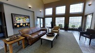 This single-floor space features a thoughtfully designed lobby lounge that immediately impresses with its dramatic vaulted ceilings and an expansive wall of large, multi-pane windows. The generous glazing draws in abundant natural light while offering clear views of the surrounding outdoor greenery, creating a bright and airy environment throughout the day.

The seating area is anchored by a rich brown leather sofa paired with a matching tan upholstered armchair, arranged around a sleek white coffee table accented by a woven bowl centerpiece. A wooden side table complements the warm tones of the furnishings, adding to the inviting character of the space.

The walls are finished in a warm taupe tone that complements the neutral carpeting, while tile flooring defines the transitional areas near the entrance. Potted plants are strategically placed throughout, adding life and a touch of natural texture to the interior. A framed wall-mounted display, resembling an informational or artistic panel, adds visual interest to one accent wall.

Wall-mounted lighting fixtures provide subtle ambient illumination, supplementing the natural light. The overall design balances comfort with a professional aesthetic, making the space well-suited for welcoming guests and facilitating relaxed, informal interactions.
