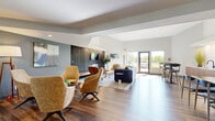 Located at 6400 Sycamore Ln N in Maple Grove, Minnesota, this single-floor communal space offers a thoughtfully designed interior that blends modern architecture with mid-century modern aesthetics. The open-concept layout flows seamlessly from a welcoming hallway into a spacious living room, unified by rich hardwood-style flooring throughout.

The living room serves as the centerpiece of the space, anchored by a dramatic dark wood-paneled accent wall housing a sleek linear fireplace and a wall-mounted television. Soaring ceilings with angled architectural details amplify the sense of openness, while large windows and glass doors flood the interior with natural light and offer views of the surrounding outdoor landscape. The lounge area is arranged with plush mid-century swivel chairs in warm amber tones, a navy sectional sofa, and accent furnishings that create multiple inviting conversation zones.

A dedicated dining and bar area adjoins the main lounge, featuring high-top tables with bar seating, a built-in banquette with teal upholstery, and a long communal dining table — all beneath pendant lighting and recessed ceiling fixtures. Subtle cool-toned wall colors contrast warmly with the wood tones and mustard accents present throughout, creating a cohesive and welcoming ambiance well-suited for both casual gatherings and relaxed lounging.