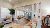 This single-floor space in Shreveport, Louisiana offers an open-concept layout that seamlessly connects the living room and kitchen into one cohesive, functional area. The living room is defined by striking architectural details, including tall white decorative columns, coffered ceilings with crown molding, and rich hardwood flooring throughout. Large windows with horizontal blinds invite abundant natural light, enhancing the warm, neutral color palette of the walls. A classic white-mantled fireplace with a decorative tile surround serves as a focal point, topped with a large-scale abstract painting that adds a contemporary artistic touch. A built-in shelving unit with an integrated media center provides both style and practicality, while ceiling fans and recessed lighting ensure comfortable illumination throughout. The kitchen features white cabinetry, granite countertops, and a vibrant teal accent wall that lends the space a lively, distinctive character. Light ceramic tile flooring, a breakfast bar with barstool seating, and stainless steel appliances complete the kitchen's functional and appealing design. Together, these spaces create a bright, elegant, and well-designed environment suited for both everyday use and social gatherings.