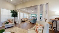 This single-floor space in Shreveport, Louisiana offers an open-concept layout that seamlessly connects the living room and kitchen into one cohesive, functional area. The living room is defined by striking architectural details, including tall white decorative columns, coffered ceilings with crown molding, and rich hardwood flooring throughout. Large windows with horizontal blinds invite abundant natural light, enhancing the warm, neutral color palette of the walls. A classic white-mantled fireplace with a decorative tile surround serves as a focal point, topped with a large-scale abstract painting that adds a contemporary artistic touch. A built-in shelving unit with an integrated media center provides both style and practicality, while ceiling fans and recessed lighting ensure comfortable illumination throughout. The kitchen features white cabinetry, granite countertops, and a vibrant teal accent wall that lends the space a lively, distinctive character. Light ceramic tile flooring, a breakfast bar with barstool seating, and stainless steel appliances complete the kitchen's functional and appealing design. Together, these spaces create a bright, elegant, and well-designed environment suited for both everyday use and social gatherings.