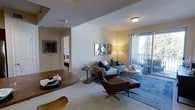 This single-floor residence showcases an open-concept layout that seamlessly connects the dining and living areas, creating a cohesive and functional living space. The dining area features a warm wooden table, providing an inviting spot for meals, while the adjacent living room is anchored by a plush sofa accented with decorative pillows in blue and yellow tones. A rich brown leather accent chair with an ottoman adds a sophisticated focal point to the seating arrangement.

The living room walls are adorned with a curated gallery of framed artwork, contributing to the space's polished aesthetic. Ambient lighting from a floor lamp casts a warm glow throughout the room, complementing the neutral wall tones and light-colored carpeting underfoot. A blue area rug defines the seating zone, adding depth and color contrast.

Natural light flows generously into the living area through a sliding glass door that opens to a private balcony, offering outdoor views and ventilation. A glimpse through an interior doorway reveals a bedroom beyond, reinforcing the thoughtful, connected flow of the floor plan. The overall ambiance is warm, modern, and well-appointed.