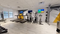Located at 507 E Main St in Bound Brook, NJ, this modern fitness facility offers a thoughtfully designed single-floor workout space that balances functionality with a sleek aesthetic. The interior features light wood-style flooring throughout, a drop-panel ceiling with recessed lighting, and a crisp white and grey color palette accented by bold yellow and dark graphite geometric wall details that give the space a dynamic, energetic character.

The cardio area is well-stocked with multiple Matrix treadmills, elliptical cross-trainers, and spin bikes, all positioned along a wall lined with flat-screen televisions to keep members engaged during workouts. Full-length mirrors amplify the sense of space and allow users to monitor their form. The strength training zone features a cable pulley machine, adjustable weight benches, and a rack of free weights, all neatly arranged to maximize usable floor space.

A transitional corridor connects the main workout areas and provides access to private rooms, including what appears to be a restroom or changing area. Sanitization stations are conveniently placed near entry points, reflecting a focus on cleanliness and member wellness. Natural light filters in through windows in the strength training area, complementing the bright, well-ventilated atmosphere throughout the facility.