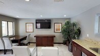 This single-floor space presents a well-organized and functional interior designed to welcome visitors with a clean, professional atmosphere. The main area features light-toned tile flooring that extends throughout, complementing the neutral gray walls and creating a bright, open feel. Recessed lighting and a textured ceiling add depth and ensure the space is well-lit throughout.

A focal point of the room is the wall-mounted flat-screen television, flanked by two framed artworks with warm-toned prints in gilded frames, lending a polished aesthetic to the space. Beneath the TV sits a rich, dark-wood credenza-style counter that serves as a functional centerpiece, ideal for reception or display purposes. White upholstered chairs are arranged to provide comfortable seating for visitors, while an additional table and chair arrangement along one side accommodates further use.

Adding a natural and inviting element, large, lush indoor plants — including a striking tall palm-like specimen — are clustered in one corner, bringing vibrancy and life to the space. To one side, cabinetry with granite-style countertops provides additional storage and utility. The overall ambiance is clean, professional, and welcoming, reflecting a space thoughtfully designed for comfort and functionality.