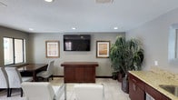 This single-floor space presents a well-organized and functional interior designed to welcome visitors with a clean, professional atmosphere. The main area features light-toned tile flooring that extends throughout, complementing the neutral gray walls and creating a bright, open feel. Recessed lighting and a textured ceiling add depth and ensure the space is well-lit throughout.

A focal point of the room is the wall-mounted flat-screen television, flanked by two framed artworks with warm-toned prints in gilded frames, lending a polished aesthetic to the space. Beneath the TV sits a rich, dark-wood credenza-style counter that serves as a functional centerpiece, ideal for reception or display purposes. White upholstered chairs are arranged to provide comfortable seating for visitors, while an additional table and chair arrangement along one side accommodates further use.

Adding a natural and inviting element, large, lush indoor plants — including a striking tall palm-like specimen — are clustered in one corner, bringing vibrancy and life to the space. To one side, cabinetry with granite-style countertops provides additional storage and utility. The overall ambiance is clean, professional, and welcoming, reflecting a space thoughtfully designed for comfort and functionality.