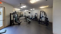 Located at 4050 Rocky Cir in Tampa, Florida, this fitness center offers a comprehensive workout facility designed to accommodate a wide range of exercise needs. The space is laid out across a single floor and is divided into distinct functional zones, each dedicated to a specific type of training.

The cardio area features multiple treadmills, elliptical machines, and a stair climber, all positioned in front of large wall-to-wall mirrors that visually expand the space and allow users to monitor their form. Ceiling fans and overhead lighting keep the area well-ventilated and bright.

The strength training section is generously equipped with a multi-station cable machine, a Smith machine with a full rack of weight plates, an adjustable bench, and a dedicated free weights area housing a tiered dumbbell rack with a full range of dumbbells. A stability ball and additional benches round out this section.

A spin bike station provides an option for high-intensity cycling workouts, positioned near decorative wall art that adds a touch of personality to the space. A vending machine offering refreshments is conveniently accessible near the entrance.

Throughout, the facility features dark rubber flooring for safety and durability, a textured ceiling, and wall-mounted mirrors and televisions. Arched doorways and soft neutral wall tones contribute to a clean, functional aesthetic throughout the entire space.