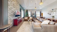 Located in Noblesville, Indiana, this thoughtfully designed community gathering space seamlessly blends a living room and dining area into one open, welcoming floor plan. The living area is anchored by a striking floor-to-ceiling stacked stone accent wall, which serves as a dramatic backdrop for the mounted flat-screen television and generous sectional seating. A tray ceiling with pendant lighting adds architectural interest overhead, while plush carpeting underfoot contributes to the room's warm, comfortable ambiance. Bold teal accent walls provide vibrant color contrast against the neutral tones throughout.

Large windows on multiple walls flood the interior with natural light and offer pleasant views of the outdoor grounds and pool area. The dining section features several round tables with mid-century modern style chairs, creating an inviting setting for communal meals or casual gatherings. A built-in window bench with cushions along one wall offers additional seating and a cozy nook to enjoy the natural light. The space also connects to a kitchen area visible in the background, complete with bar-height seating. Every corner of this community room reflects a balance of functionality and style, making it an ideal space for socializing and relaxation.