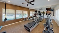 This single-floor amenity space is designed to support an active lifestyle, featuring two distinct areas: a fully equipped fitness center and a resort-style outdoor pool. The fitness center is thoughtfully appointed with multiple treadmills, a dumbbell rack, and additional cardio equipment. A large wall-to-wall mirror expands the sense of space, while a ceiling fan and recessed lighting ensure a comfortable workout environment. Multiple windows fitted with blinds flood the room with natural light and offer views of the surrounding greenery. A wall-mounted television provides entertainment during workouts, and a workout chart is conveniently posted for reference. The carpeted flooring adds a layer of comfort underfoot. The outdoor pool area is equally impressive, featuring a generously sized swimming pool with clear turquoise water, framed by a wrought-iron fence for safety. Lounge chairs are positioned poolside for relaxation. Tall palm trees and mature landscaping surround the area, creating a tranquil, resort-like atmosphere beneath open blue skies. Together, these amenities provide a comprehensive recreational offering in a well-maintained, inviting setting.