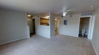 This single-floor residential unit offers a clean, functional layout designed for comfortable everyday living. The main living area is expansive and open, finished with neutral wall-to-wall carpeting that lends a warm, understated feel to the space. A ceiling fan is centrally positioned overhead, providing both light and airflow to the room.

A half-wall partition separates the living area from the kitchen, featuring a pass-through counter that promotes an open, connected feel between the two spaces. The kitchen is equipped with warm wood-tone cabinetry and overhead lighting, providing ample storage and workspace. A white refrigerator is visible toward the rear of the kitchen, and the space is efficiently arranged for practical use.

Adjacent to the entry, a utility or electrical panel is mounted on the wall, reflecting the unit's straightforward and functional design. A doorway near the entry leads to additional rooms, and a corridor visible beyond the kitchen suggests access to a bathroom or further living quarters. The unit's neutral color palette of soft whites and light grays creates a bright, cohesive atmosphere throughout. Ventilation registers are visible in the ceiling, ensuring consistent climate control. Overall, the space presents a well-organized, move-in-ready layout suited for a range of residents.