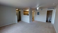 This single-floor residential unit offers a clean, functional layout designed for comfortable everyday living. The main living area is expansive and open, finished with neutral wall-to-wall carpeting that lends a warm, understated feel to the space. A ceiling fan is centrally positioned overhead, providing both light and airflow to the room.

A half-wall partition separates the living area from the kitchen, featuring a pass-through counter that promotes an open, connected feel between the two spaces. The kitchen is equipped with warm wood-tone cabinetry and overhead lighting, providing ample storage and workspace. A white refrigerator is visible toward the rear of the kitchen, and the space is efficiently arranged for practical use.

Adjacent to the entry, a utility or electrical panel is mounted on the wall, reflecting the unit's straightforward and functional design. A doorway near the entry leads to additional rooms, and a corridor visible beyond the kitchen suggests access to a bathroom or further living quarters. The unit's neutral color palette of soft whites and light grays creates a bright, cohesive atmosphere throughout. Ventilation registers are visible in the ceiling, ensuring consistent climate control. Overall, the space presents a well-organized, move-in-ready layout suited for a range of residents.