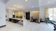 This single-floor space showcases a refined contemporary design with an open-concept layout that seamlessly connects the kitchen and dining areas. The kitchen is anchored by a substantial island finished in a speckled stone surface, accompanied by stylish black bar stools that invite casual seating. Pendant lights with woven shades hang above the island, complementing the recessed lighting throughout the space. Dark cabinetry contrasts elegantly with the light-toned tile flooring that runs throughout, while stainless steel appliances add a modern, functional touch. A textured accent wall in warm tones adds visual depth near the kitchen. A standout feature is the intimate built-in dining booth, nestled within a defined alcove and framed by a striking three-dimensional wave-textured wall panel with accent lighting, creating a cozy yet sophisticated atmosphere. A bold dark wood entry door serves as a strong focal point, grounding the open space. The overall ambiance is clean, curated, and modern, with a careful balance of light and dark elements throughout.