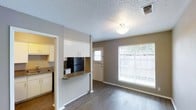 This single-floor space offers a practical and well-organized layout designed for comfortable everyday living. The open-concept design connects the kitchen and living area seamlessly, anchored by a half-wall pass-through that adds both functionality and a sense of openness. The kitchen is equipped with white cabinetry, a black refrigerator, and a dark countertop that extends to form the pass-through bar, offering additional prep and casual dining space. A sink and dishwasher are also visible, completing the kitchen's functional setup. Overhead cabinetry above the pass-through provides extra storage while maintaining visual connectivity between the spaces. The living area is finished with warm hardwood-style flooring that runs throughout, adding a cohesive and modern touch to the interior. A large window floods the living space with natural light, creating a bright and welcoming ambiance, while a secondary window near the entryway enhances ventilation and daylight. The textured ceiling adds a classic finishing detail throughout. The front entry door opens directly into the living space, offering a straightforward and accessible layout. Overall, the space presents a clean, neutral palette that provides a versatile canvas for a variety of interior styles.