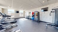 Located in Box Elder, South Dakota, this single-floor fitness center is designed to support a comprehensive workout experience. The space is organized into distinct training zones, each outfitted with purpose-specific equipment. The cardio area features a row of treadmills, elliptical trainers, a stationary bike, and rowing machines, all positioned beneath wall-mounted flat-screen TVs for entertainment during workouts. Large windows line the walls throughout, flooding the facility with natural light and offering open views of the surrounding landscape. The free weight zone includes a full dumbbell rack spanning a wide range of weights, alongside kettlebells, medicine balls, and exercise balls neatly stored on a dedicated rack. A cable resistance machine anchors the strength training section, complemented by large wall mirrors that enhance the sense of space and allow for form monitoring. The flooring throughout is a durable dark rubberized surface well-suited for high-activity use. The drop ceiling with recessed and flush lighting ensures consistent illumination across all areas. The overall design is clean, modern, and functional, with neutral tones and a bright, open atmosphere that makes the space welcoming and practical for users of all fitness levels.