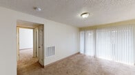 This single-floor residential unit offers a functional and comfortable layout with a clean, neutral aesthetic throughout. The spacious living area is finished with plush wall-to-wall carpeting and features large sliding glass doors dressed with vertical blinds, allowing abundant natural light to fill the room. A ceiling-mounted light fixture and a textured popcorn ceiling complete the space. An adjacent dining or secondary living area is highlighted by an elegant chandelier and similarly large window treatments, reinforcing the bright and open feel of the unit.

The kitchen is well-appointed with warm wood cabinetry, light granite-style countertops, a black gas range with an overhead exhaust hood, and a sink — providing a practical and attractive cooking environment with hardwood-style flooring underfoot.

The bedroom areas are generously sized, each with soft carpeting, vertical blinds for natural light control, and access to their own bathrooms. Two bathrooms are visible from a central hallway, each featuring warm wood vanity cabinetry, mirrors with overhead lighting, and hardwood-style flooring — offering a well-organized and private layout. The overall floor plan, as shown in the dollhouse view, reveals a thoughtfully arranged interior with clear separation between living, sleeping, and utility zones.