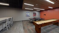 Located at 29 E Green St in Champaign, Illinois, this single-floor amenity space is thoughtfully designed to offer a dynamic and social environment. The space is unified by a consistent design language featuring warm wood slat ceilings, recessed linear lighting, and a neutral color palette accented by bold orange walls that add energy throughout.

The game room serves as a central hub for recreation, equipped with a full-size ping-pong table and a classic shuffleboard table. Colorful framed artwork decorates the walls, and patterned carpet tiles with orange accents complement the room's vibrant aesthetic.

Adjacent to the game area is a comfortable lounge space furnished with modular blue seating, a coffee table, and multiple wall-mounted flat-screen TVs, making it well-suited for both casual gatherings and screen-based entertainment. The wood slat ceiling treatment continues here, lending warmth and cohesion to the space.

The coffee bar area offers a clean, modern design with white cabinetry, a dark countertop, a utility sink, and a built-in coffee machine. A concrete-finish high-top table with bar-height seating provides a spot for casual conversation. Decorative coffee bean artwork lines the wall, reinforcing the space's café-inspired character. Large-format tile flooring and a dramatic dark ceiling complete the polished, contemporary look.