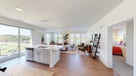This single-floor residence is designed with an open-concept layout that seamlessly connects the kitchen, dining, and living areas, creating a cohesive and functional living space. The kitchen is outfitted with grey shaker-style cabinetry, white quartz countertops, stainless steel appliances, a subway tile backsplash, and a large island with bar seating. A sliding glass door provides direct outdoor access and fills the space with natural light. The dining area features a bright corner setting with multiple windows offering outdoor views, making it an inviting space for everyday meals. The expansive living room benefits from a generous bank of windows, ensuring ample natural light, and opens to a dedicated home office nook with built-in shelving. The primary bedroom offers a serene retreat with a bold botanical accent wall and large windows, while a second bedroom is finished with neutral tones and natural light. The bathroom includes a wide vanity with ample storage, wood-look flooring, and a glass-enclosed shower. Throughout, warm wood-look flooring, crisp white walls, and contemporary finishes maintain a clean, modern aesthetic.