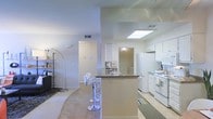 This single-story space features an open-concept design that integrates the kitchen and living areas into one cohesive layout. The kitchen is equipped with white cabinetry, white appliances including a refrigerator and range, and an under-cabinet range hood. A peninsula-style kitchen island with a dark countertop serves as a functional workspace and casual dining area, accompanied by modern chrome bar stools. A skylight above the kitchen fills the space with natural light, complementing the neutral wall tones and light tile flooring throughout. The living area transitions smoothly from the kitchen, featuring a area rug that defines the space, and a sleek arc floor lamp that adds ambient lighting. Visible hallways lead to additional rooms, suggesting a well-connected floor plan. HVAC vents are positioned throughout the ceiling, ensuring consistent climate control. The overall ambiance is bright, clean, and functional, with a neutral color palette of whites and warm beiges that creates an inviting and versatile environment suitable for a variety of uses.