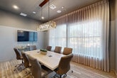 Located at 2926 Barker Cypress Rd in Houston, TX, this professional conference room offers a refined and functional setting well-suited for meetings and collaborative work. The room is anchored by a large, light-wood rectangular conference table surrounded by cushioned, high-back brown leather rolling chairs, comfortably seating up to eight participants. A striking geometric pendant chandelier hangs centrally above the table, providing warm, focused illumination that complements the recessed ceiling lights throughout the space. A large wall-mounted flat-screen television is positioned at one end of the room, offering seamless presentation capabilities, with a sleek credenza nearby for additional equipment or storage. Floor-to-ceiling sheer drapery lines an expansive window wall, filtering abundant natural light while maintaining a sense of privacy and softening the overall ambiance. The flooring features a multi-toned carpet tile that adds texture and a contemporary touch. A distinctive wood-framed glass partition wall with transom windows connects the room to an adjacent common area, providing visual openness while maintaining acoustic separation. Tasteful artwork and subtle greenery complete the space, contributing to a calm, professional atmosphere ideal for business gatherings.