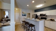 This single-floor residence offers two bedrooms and two bathrooms within a thoughtfully designed open-concept layout. The kitchen serves as a central highlight, featuring white cabinetry that extends to the upper walls, a stainless steel refrigerator, built-in microwave, and a range. A bold navy accent wall adds a contemporary contrast to the otherwise neutral color palette throughout the space. The kitchen flows seamlessly into the living area via a peninsula-style breakfast bar equipped with a grey countertop and seating for three, making it ideal for casual dining or entertaining. Recessed and flush-mount ceiling fixtures provide consistent, well-distributed lighting throughout. The entryway, finished with tile flooring, offers a clean transition into the main living space, which features plush carpeting. Neutral-toned walls are complemented by decorative artwork, contributing to a welcoming ambiance. A textured ceiling adds subtle dimension to the interior. The open sightlines between the kitchen, dining, and entry areas create a connected, functional living environment well-suited for everyday comfort.