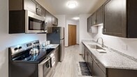 This single-floor unit offers a thoughtfully designed layout that balances comfort and functionality. The galley-style kitchen stands out as a highlight, featuring dark taupe cabinetry, granite-look countertops, stainless steel appliances including a refrigerator, electric range, and microwave, and light wood-look luxury vinyl flooring — creating a clean, modern aesthetic.

The hallway connects the main living areas seamlessly, with wood-framed doorways providing a warm contrast to the crisp white walls. The bathroom includes a vanity with matching dark cabinetry and granite-look countertops, complementing the kitchen's finishes for a cohesive look throughout.

The bedrooms are spacious and carpeted, each equipped with a wall-mounted air conditioning unit and baseboard heating for year-round climate control. Windows dressed with horizontal blinds allow natural light to filter in. Each bedroom is served by generous closet space — one featuring a large walk-in closet with dual wire shelving, and another with a full-width wire shelf and hanging rod, providing ample storage.

Throughout the unit, warm wood-toned baseboards and door trims add character, while textured ceilings and flush-mount lighting provide a clean, finished look. The overall ambiance is tidy, neutral, and move-in ready.