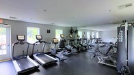 Located at 2 Dutton Drive in Matawan, NJ, this single-floor fitness center offers a clean, functional environment designed to support a comprehensive workout experience. The space is outfitted with a variety of modern cardio equipment, including treadmills, elliptical trainers, and rowing machines, all arranged to maximize usability and floor space. Large floor-to-ceiling mirrors line one wall, creating a sense of expanded space while allowing users to monitor their form during exercise. Industrial-style standing fans are positioned throughout the room, ensuring proper airflow and comfort during workouts. The dark rubberized flooring provides both durability and safety underfoot, while recessed ceiling lighting keeps the space evenly illuminated. Multiple windows along the exterior walls invite generous natural light and offer views of the surrounding greenery, contributing to an open and energizing atmosphere. The walls are finished in a neutral light gray tone, lending the space a modern and professional aesthetic. Overall, the fitness center presents a well-maintained, practical facility suited for a range of cardio and general fitness activities.