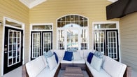 This inviting covered outdoor patio showcases a classic Southern-inspired architectural design, highlighted by warm yellow horizontal lap siding, crisp white trim, and stately white columns resting on stone bases. The space is thoughtfully organized into multiple seating areas, each furnished with all-weather wicker sectionals and chairs dressed in white cushions and navy blue accent pillows, offering comfortable spots to gather and unwind. A central focal point of the patio is a striking arched window flanked by large French doors with black frames, offering clear views through to a pool area beyond. Recessed lighting and a ceiling fan are visible within the arched ceiling detail, adding both function and charm. An outdoor wall-mounted television is positioned along one side of the patio, making it well-suited for casual outdoor entertainment. The covered wraparound design provides generous shade across the polished concrete floor, while open columns allow for natural breezes and natural light to filter through. The verdant surroundings of mature trees and manicured landscaping are visible from the porch, enhancing the overall sense of a serene and well-appointed outdoor living environment. Wall-mounted lantern-style light fixtures complete the refined yet relaxed aesthetic of this exceptional outdoor space.