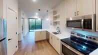 Located on NE Main St in Minneapolis, this single-floor unit showcases a clean, contemporary design highlighted by light wood-tone cabinetry, white quartz countertops, and warm hardwood-style flooring throughout. The open kitchen is thoughtfully appointed with stainless steel appliances, including a refrigerator, electric range, built-in microwave, and dishwasher. White subway tile backsplash, a deep undermount sink with a matte black faucet, and a mix of upper cabinets and open shelving complete the well-organized space. Pendant lighting above the kitchen peninsula adds a stylish focal point, while recessed lighting ensures consistent illumination. A full-height glass sliding door opens directly to a private balcony, flooding the living area with natural light and offering leafy outdoor views. The bedroom is generously proportioned, featuring a ceiling fan, large black-framed windows with tree-lined views, and durable hardwood-style flooring. The bathroom is finished with white subway tile in the tub surround, large-format floor tiles, a light wood vanity with a white countertop, a matte black faucet, and a bold dark accent wall. A stacked in-unit washer and dryer, along with open built-in shelving for additional storage, are conveniently integrated into the bathroom space.