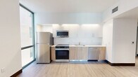 Located at 224 23rd St in Miami Beach, FL, this modern unit offers four bedrooms and one bathroom across a single floor. The interior is defined by a clean, contemporary aesthetic with light wood-look flooring, crisp white walls, and dark contrasting baseboards running throughout. Large floor-to-ceiling windows flood the living and bedroom areas with natural light and provide views of the surrounding neighborhood. The open kitchen is well-appointed with two-tone cabinetry — white upper cabinets paired with natural wood-finish lower cabinets — and a full suite of stainless steel appliances including a refrigerator, electric range, over-the-range microwave, and dishwasher, complemented by light countertops and recessed under-cabinet lighting. The bedrooms are compact yet functional, with select rooms featuring dedicated closet space and access to natural light. The shared bathroom is finished with white subway tile surrounding a full soaking tub and shower, a wood-vanity with a white countertop, a large frameless mirror, and wood-look tile flooring. Recessed and flush-mount ceiling lighting is found throughout, contributing to a bright, well-lit environment across all rooms.