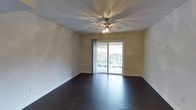 This single-floor space offers a clean and functional layout with a comfortable, straightforward design. The main living area is defined by rich, dark hardwood-style flooring that runs throughout, providing a striking contrast against the light-toned walls. A textured ceiling adds subtle visual interest overhead, while a multi-blade ceiling fan with an integrated light fixture serves as a central functional feature, ensuring air circulation and ambient lighting throughout the room. One of the standout elements of this space is the large sliding glass door, which floods the interior with generous natural light and provides direct access to a private balcony enclosed by a metal railing. The outdoor view visible through the glass door suggests a pleasant surrounding environment with greenery nearby. The walls are painted in a neutral warm tone, contributing to an open and airy atmosphere. White baseboards frame the perimeter of the room, adding a clean finishing detail. The space presents a well-maintained, move-in ready environment with a practical design that balances simplicity and comfort, making it well-suited for a variety of living arrangements.