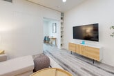 This efficiently designed one-bedroom, one-bathroom residence demonstrates how intelligent layout planning and modern aesthetics can make a compact space feel open and functional. Light wood-look flooring runs throughout, unifying the space while recessed ceiling lights provide consistent, clean illumination.

The kitchen features sleek white lower cabinetry paired with gray upper cabinets, stone-look countertops, a built-in oven, an induction cooktop, and a modern arc faucet — delivering a polished, full-function cooking space. It flows seamlessly into the living area, where a neutral sectional sofa, a round wood coffee table, and a wall-mounted flat-screen TV create a cozy yet contemporary atmosphere. Built-in shelving adds both storage and display space.

The bedroom is accessed via a frosted-glass sliding barn door and is filled with natural light from large windows with outdoor views. Elevated open shelving above the closet maximizes vertical storage. A dedicated workspace with a wood desk and chair is neatly integrated into the room.

The bathroom offers a full soaking tub with shower, light wood-tone flooring, a wall-mounted cabinet, and clean white walls — simple and functional. Throughout the residence, the design palette of whites, warm woods, and soft neutrals creates a bright, cohesive, and welcoming ambiance.
