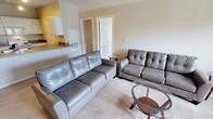 This single-floor residence offers a comfortable and functional layout designed for everyday living. The living room serves as the central gathering space, featuring wall-to-wall carpeting that adds warmth underfoot. The room is furnished with a full-sized grey tufted leather sofa and a matching loveseat, arranged around a round glass-top coffee table, creating an inviting seating arrangement. A smaller glass side table accents the space between the two seating pieces. The neutral, warm-toned walls complement the light carpeting and contribute to a calm, understated ambiance throughout. Adjacent to the living area, the kitchen is visible through an open pass-through layout, showcasing white upper cabinetry and rich stone countertops that provide a subtle contrast. The kitchen space appears bright and efficiently organized. Interior doors with classic panel detailing provide access to additional rooms within the residence, suggesting a well-connected floor plan. The overall design emphasizes practicality and comfort, with a cohesive neutral color palette running throughout the space that makes it feel clean, open, and move-in ready.