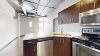 This single-floor urban unit showcases a distinctive industrial-chic aesthetic that blends raw architectural elements with modern finishes throughout its six rooms. Polished concrete floors run continuously across the space, lending a sleek, contemporary character that ties each room together cohesively.

The kitchen is a standout feature, outfitted with rich dark-wood cabinetry, light-toned countertops, and a full suite of stainless steel appliances including a refrigerator, electric range, over-the-range microwave, and dishwasher. A peninsula with additional cabinetry provides extra prep space and storage.

The open living area benefits from large windows that draw in generous natural light and offer views to an outdoor balcony or walkway. The exposed ceiling features industrial-style ductwork, conduit, and pendant lighting fixtures, reinforcing the urban loft aesthetic.

The bathroom continues the polished concrete flooring and includes a wide floating vanity with an undermount sink, a large mirror, pendant lighting, and a full bathtub with shower. The bedroom offers a clean, minimalist layout with a mirrored sliding-door closet and additional natural light from a tall window. The crisp white walls throughout create a bright, airy ambiance that complements the bold industrial details.