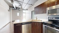 This single-floor urban unit showcases a distinctive industrial-chic aesthetic that blends raw architectural elements with modern finishes throughout its six rooms. Polished concrete floors run continuously across the space, lending a sleek, contemporary character that ties each room together cohesively.

The kitchen is a standout feature, outfitted with rich dark-wood cabinetry, light-toned countertops, and a full suite of stainless steel appliances including a refrigerator, electric range, over-the-range microwave, and dishwasher. A peninsula with additional cabinetry provides extra prep space and storage.

The open living area benefits from large windows that draw in generous natural light and offer views to an outdoor balcony or walkway. The exposed ceiling features industrial-style ductwork, conduit, and pendant lighting fixtures, reinforcing the urban loft aesthetic.

The bathroom continues the polished concrete flooring and includes a wide floating vanity with an undermount sink, a large mirror, pendant lighting, and a full bathtub with shower. The bedroom offers a clean, minimalist layout with a mirrored sliding-door closet and additional natural light from a tall window. The crisp white walls throughout create a bright, airy ambiance that complements the bold industrial details.