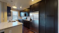 Located at 14-20 E Hubbard Ave in Columbus, OH, this single-floor residence offers a well-designed layout with a blend of modern finishes and functional living spaces. The galley-style kitchen is a standout feature, showcasing rich dark wood cabinetry extending floor to ceiling, light granite countertops, a subway tile backsplash, and a full suite of stainless steel appliances including a refrigerator with ice maker, electric range, and built-in microwave. Pendant lighting and a window overlooking the surroundings add warmth and natural light to the space. The open living area is characterized by gleaming dark hardwood floors, crown molding, recessed lighting, and an abundance of natural light streaming through large windows. A door provides direct access to a private balcony, extending the living space outdoors. The bedrooms vary in finish — one features rich hardwood floors and large windows with city views, while another is carpeted for added comfort. Bathrooms are well-appointed, with one full bath featuring tile flooring, a soaking tub with tiled surround, dark vanity cabinetry, and a quartz countertop. An in-unit laundry area adds everyday convenience. Throughout, warm neutral tones and consistent finishes create a cohesive and inviting atmosphere.