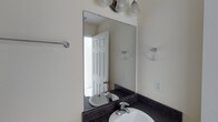 This single-story space includes a well-appointed bathroom that demonstrates a practical yet polished design. The vanity area serves as a focal point, showcasing a dark speckled granite countertop that contrasts cleanly against the light, neutral-toned walls. An undermount white sink is paired with a chrome two-handle faucet, contributing to a cohesive and timeless aesthetic. A large frameless mirror is mounted above the vanity and is illuminated by a multi-bulb vanity light fixture in a matching chrome finish, ensuring ample brightness for daily use. Chrome towel rings are positioned on either side of the mirror, offering convenient accessibility, while a chrome towel bar is also present, adding to the bathroom's functional layout. A standard white six-panel door provides entry to the space, and a traditional outlet is installed along the wall. The overall color palette — composed of warm off-white walls, dark countertops, and polished chrome hardware — creates a clean, neutral environment that is both inviting and easy to maintain.