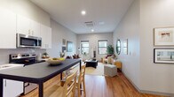 This single-floor residence is designed with a clean, contemporary aesthetic that emphasizes open living and functional space. The layout flows seamlessly from the kitchen into the living area, creating a cohesive and welcoming environment throughout.

The kitchen is outfitted with white cabinetry extending to the ceiling, stainless steel appliances including a refrigerator, range, and microwave, and a dark-topped breakfast bar with seating for two. Track lighting illuminates the workspace effectively, while warm hardwood-style flooring runs throughout the main living areas.

The open living room receives natural light from multiple windows and offers a relaxed, residential atmosphere. A ceiling-mounted HVAC unit ensures climate comfort, and the neutral gray walls provide a versatile backdrop for furnishings.

The bedroom is a quiet retreat featuring a window with urban rooftop views, allowing natural light to fill the room. A roller shade and curtain rod offer flexible light control, and the cozy layout comfortably accommodates a full-sized bed and additional décor.

The bathroom is bright and clean, finished with light tile flooring, white cabinetry with brushed gold hardware, and a simple, uncluttered layout that prioritizes functionality and ease of use.