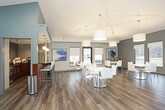 This single-floor community space is designed to serve as a welcoming gathering hub with a contemporary aesthetic throughout. The open-concept layout is immediately apparent, with rich hardwood-style flooring extending across the entire space, tying together distinct functional zones without the use of dividing walls.

The main lounge area is generously proportioned, featuring vaulted ceilings that enhance the sense of openness. Multiple seating arrangements—including a tufted leather sofa, neutral-toned armchairs, and accent tables—are organized around a round coffee table atop a vibrant teal area rug. A flat-screen television and media console anchor one end of the lounge, while large windows and glass doors invite natural light and provide views of the surrounding outdoor areas.

A dedicated dining zone accommodates smaller group gatherings with a white pedestal table and modern chairs, complemented by additional seating nearby. Large windows surround this area, maintaining the bright and airy atmosphere found throughout the space.

The kitchenette is well-appointed with warm medium-toned cabinetry, dark granite countertops, a stainless steel sink, and a full-size stainless steel refrigerator. A coffee station with supplies adds a practical hospitality touch. Gray walls throughout create a cohesive, modern backdrop that balances comfort with a polished, functional design.