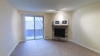 This single-floor residence offers a welcoming and functional layout designed for comfortable everyday living. The living room serves as the heart of the space, immediately drawing attention with its classic brick fireplace — a standout architectural feature that adds warmth and character to the room. The fireplace is framed by a brick surround and hearth, providing a natural focal point for the space.

Plush wall-to-wall carpeting runs throughout the living area, contributing to a cozy and inviting atmosphere. A set of sliding glass doors floods the room with natural light and provides direct access to a private balcony, offering pleasant outdoor views of the surrounding landscape. Vertical blinds frame the doors, allowing for flexible light control.

The textured ceiling adds a subtle layer of detail overhead, while neutral-toned walls provide a versatile backdrop that complements a variety of interior styles. A track light fixture above provides targeted ambient lighting. The overall design balances functional simplicity with warm, homey touches, making the space approachable and livable. The combination of natural light, outdoor access, and the brick fireplace creates an ambiance that feels both comfortable and timeless.