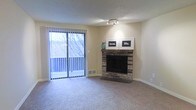 This single-floor residence offers a welcoming and functional layout designed for comfortable everyday living. The living room serves as the heart of the space, immediately drawing attention with its classic brick fireplace — a standout architectural feature that adds warmth and character to the room. The fireplace is framed by a brick surround and hearth, providing a natural focal point for the space.

Plush wall-to-wall carpeting runs throughout the living area, contributing to a cozy and inviting atmosphere. A set of sliding glass doors floods the room with natural light and provides direct access to a private balcony, offering pleasant outdoor views of the surrounding landscape. Vertical blinds frame the doors, allowing for flexible light control.

The textured ceiling adds a subtle layer of detail overhead, while neutral-toned walls provide a versatile backdrop that complements a variety of interior styles. A track light fixture above provides targeted ambient lighting. The overall design balances functional simplicity with warm, homey touches, making the space approachable and livable. The combination of natural light, outdoor access, and the brick fireplace creates an ambiance that feels both comfortable and timeless.
