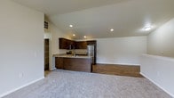 This single-floor space features an open-concept layout that seamlessly connects the kitchen and living area, creating a functional and cohesive environment. The kitchen is outfitted with rich, dark wood cabinetry that provides ample storage, complemented by granite-look countertops that add a touch of elegance. A peninsula with an integrated sink serves as both a workspace and a natural divider between the kitchen and living area, offering additional prep and seating potential. Stainless steel appliances, including a refrigerator and microwave, contribute a modern, polished look. Recessed lighting throughout the kitchen ensures bright, even illumination. The flooring transitions from hardwood-style planks in the kitchen zone to plush, neutral-toned carpet in the living area, clearly delineating the two spaces while maintaining visual harmony. The living area is generously sized with clean white walls and vaulted ceilings that enhance the sense of openness and airiness. A pantry or storage closet is visible near the kitchen, adding practical utility. The overall ambiance is clean, modern, and move-in ready.