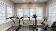 Located at 168 River in Andover, Massachusetts, this single-floor fitness center is a well-appointed exercise facility designed to support a variety of workout routines. The space is brightened by multiple large windows dressed with adjustable horizontal blinds, allowing natural light to fill the room while offering views of the surrounding property exterior. Recessed and overhead fluorescent lighting further ensures the space is consistently well-lit throughout the day. The flooring throughout is durable rubber gym-grade material, providing both safety and comfort during exercise. The cardio zone is equipped with multiple treadmills, an elliptical cross-trainer, and a recumbent stationary bike, all arranged along the windowed wall to take advantage of the natural light and outdoor views. A dedicated strength training area features a multi-function cable machine with an adjustable bench, ideal for a range of resistance exercises. The accessory corner includes a water dispenser, stability balls in multiple sizes, medicine balls, and additional small fitness tools, making the space suitable for stretching and functional training. A glass-paneled door provides a convenient entry and exit point with a hand sanitizer dispenser mounted nearby. Clean, neutral-toned walls and a practical layout give the fitness center an organized, professional atmosphere.